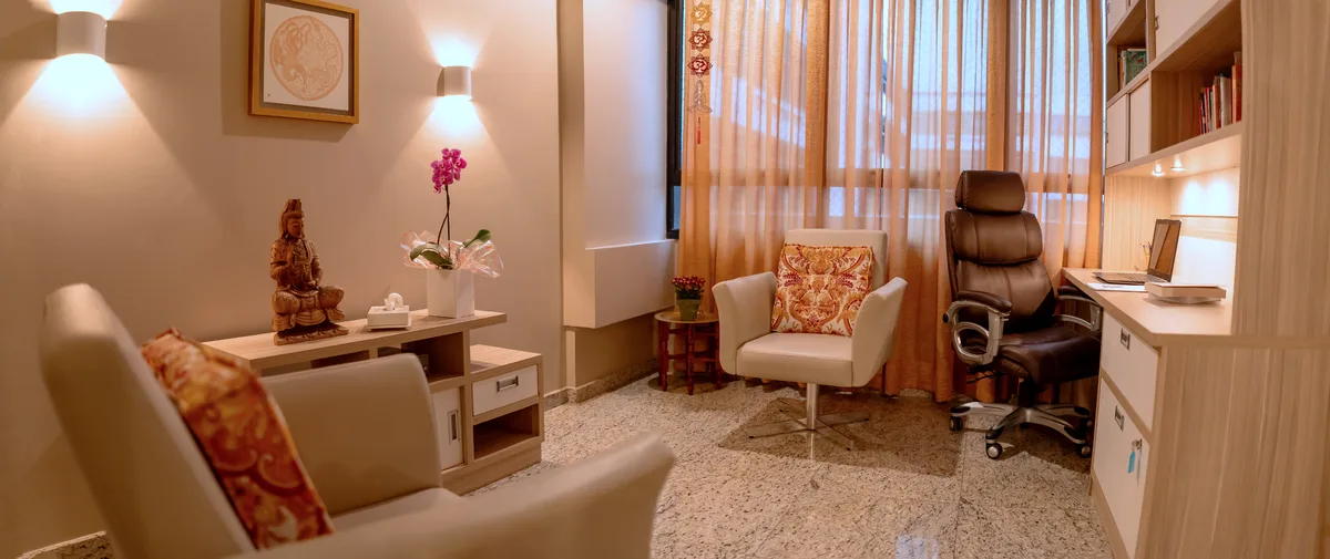 Sala de atendimento do consultório, com poltronas, estante e janela com cortina