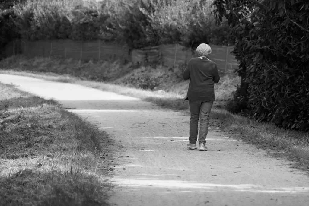 Senhora de cabelos brancos caminhando de costas por uma trilha, em preto e branco